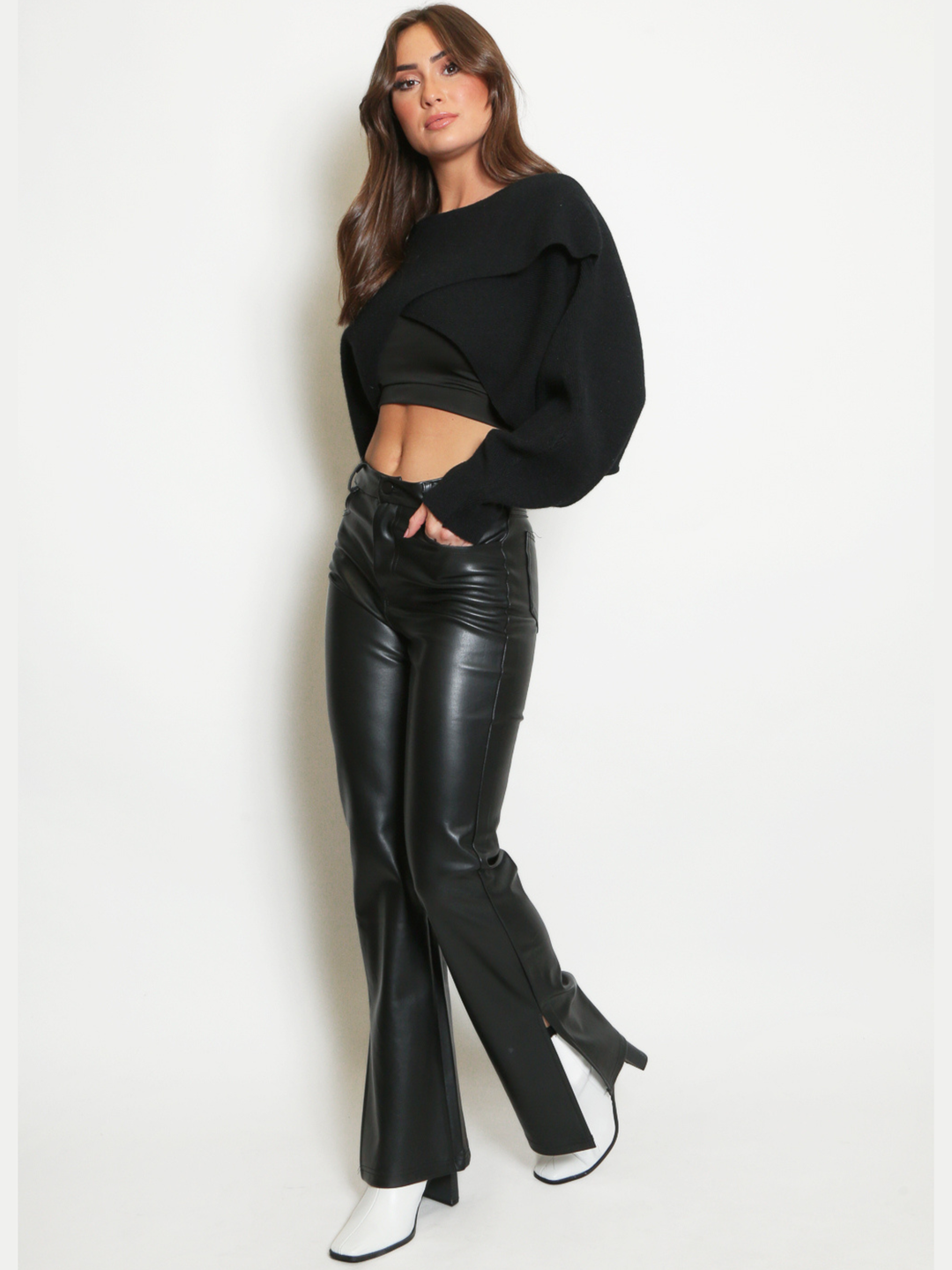 Full length shot. Female model stands in front of a plain background. The model wears a wrapped front criss cross knitted cropped jumper, with long sleeves. The model has tanned skin and long brown hair,  she stands to the side, the side of the cropped jumper is visible, showing the criss cross wrapped deign and long sleeves. The model wears pu leather trousers and side slit white boots. 