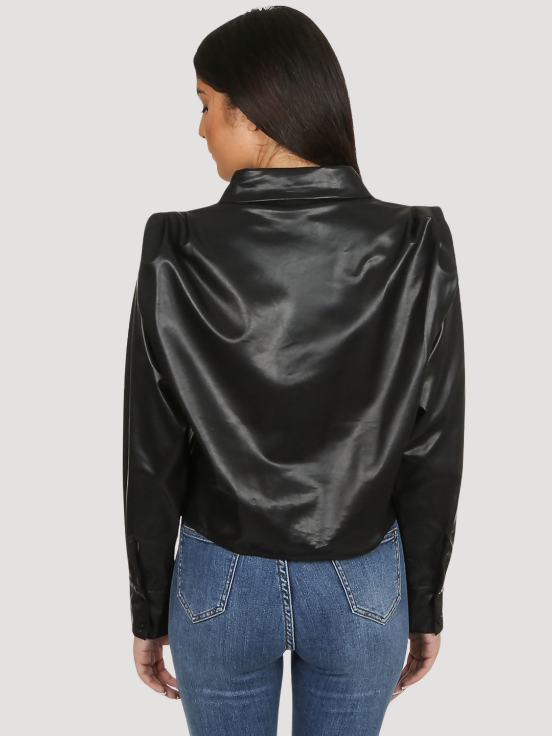 Model wears black faux leather shirt with pleated shoulders and long sleeve. The pu cropped front button shirt and collard neck. Model stands with her back to the camera. The back of the cropped pu leather shirt, the pleated puff shoulders are visible.