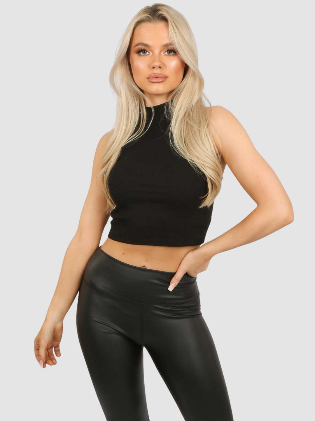 Model wears a high neck black, ribbed sleeveless jersey crop top. The model Stands in front of a plain grey background, she has long blonde hair and tanned skin, she places one hand on her hip and the other by her side. 
