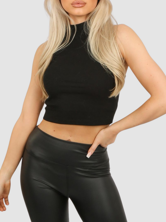 Mid length shot. Model wears a high neck black, ribbed sleeveless jersey crop top. The model Stands in front of a plain grey background, she has long blonde hair and tanned skin, she places one hand on her hip and the other by her side. 