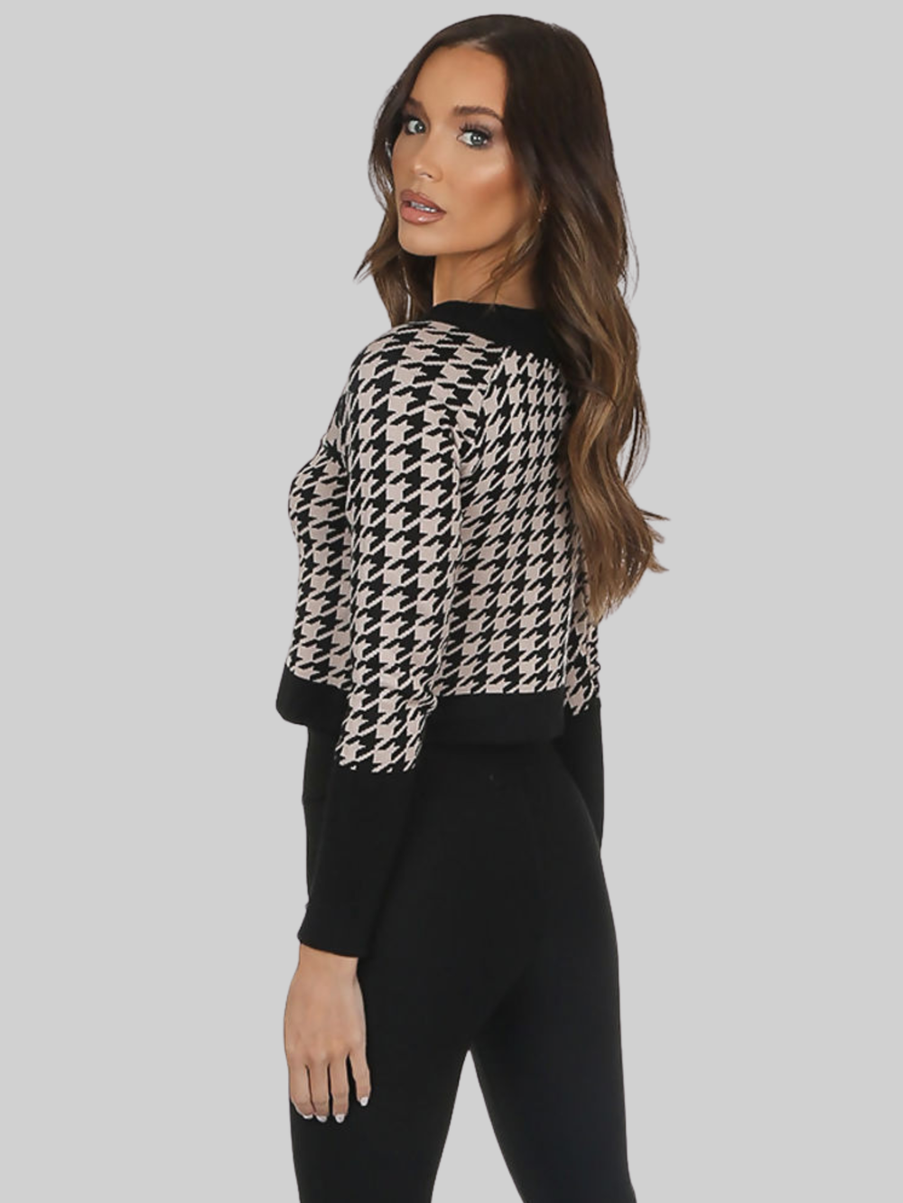 Female model stands in front of a pale grey background. The model wears a black and beige cropped cardigan with front military button fastening. The model has tanned skin and long brown hair, she stands to the side and turns her head to look directly into the camera, placing both hands by her sides. The side and back of the cropped cardigan can be seen. 