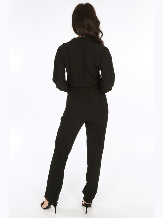 Female model wears a black jumpsuit on a white background, the black boiler suiy has front pockets and a zip front fastening as well as a belted waist tie. The model has tanned skin and long brown hair, she standswith her back to the camera, the back of the boiler suit can be seen. 