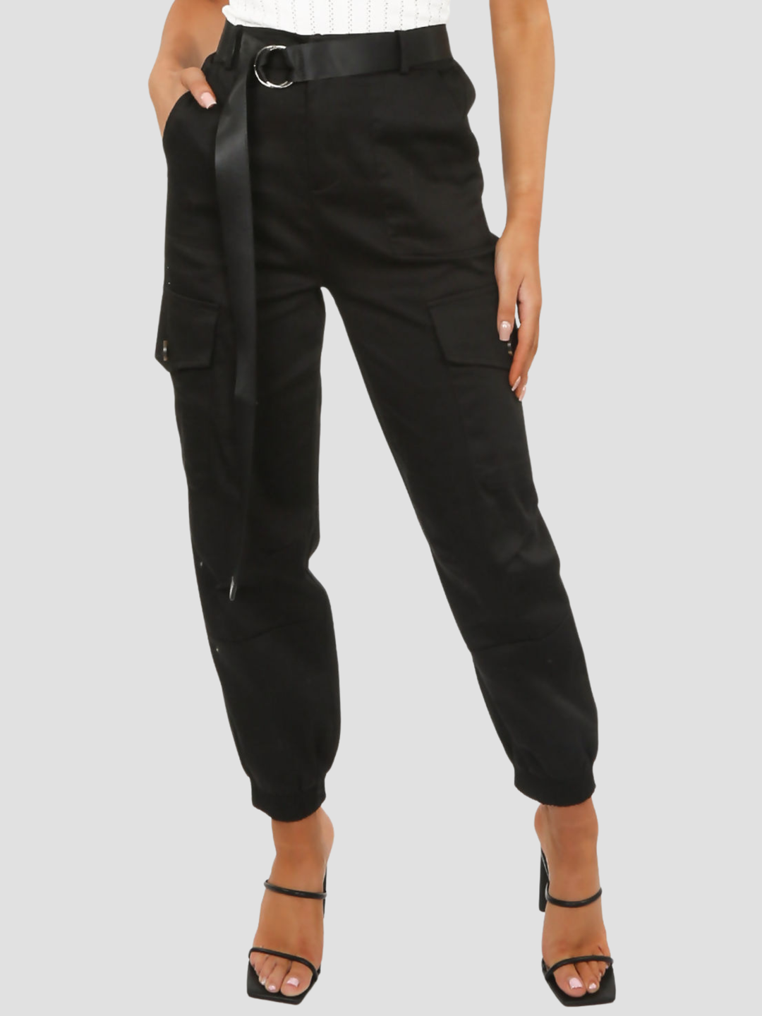 Female model stands before a plain background. The model wears a black cargo trousers with cuffed ankles and a long strap d ring belt. The model wears a high heeled shoe, she places one hand in her pocket and the other by her side. 