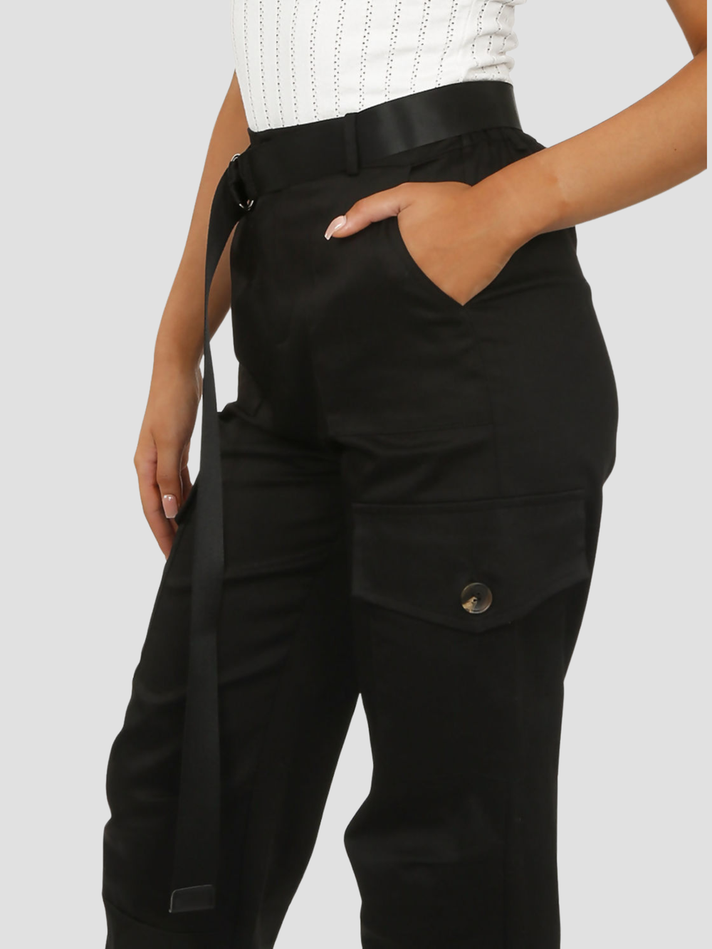 Female model stands before a plain background. The model wears a black cargo trousers with cuffed ankles and a long strap d ring belt, the model stands to the side, the side of the pocket can be seen, she places one hand in her pocket and the other by her side.