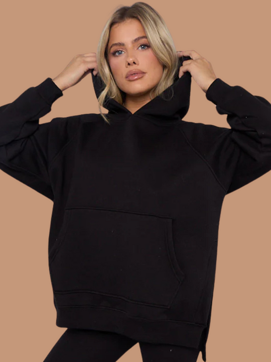 Model wears a black oversized hoodie with front functional pockets and matching  black ribbed leggings. The oversized hoodie has side splits on either side of the jumper.  Model has blonde hair,  she places both hands on the hood and looks directly into the camera.