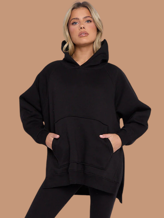 Model wears a black oversized hoodie with front functional pockets and matching  black ribbed leggings. The oversized hoodie has side splits on either side of the jumper.  Model has blonde hair, she places both hands in her pockets and faces directly into the camera.