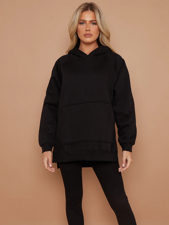 Model wears a black oversized hoodie with front functional pockets and matching  black ribbed leggings. The oversized hoodie has side splits on either side of the jumper.  Model has blonde hair, she faces forward and has both hanfs by her side.