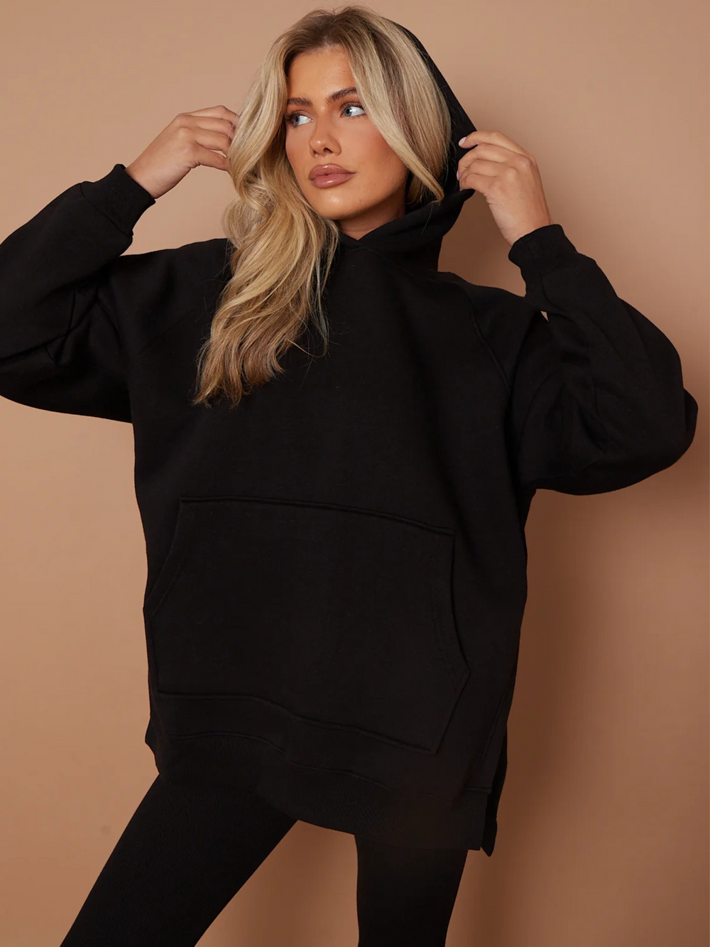 Model wears a black oversized hoodie with front functional pockets and matching  black ribbed leggings. The oversized hoodie has side splits on either side of the jumper.  Model has blonde hair, she stares to the side andholds either side of the hood.
