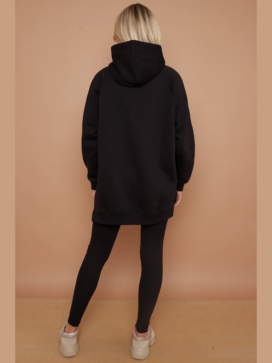 Full length shot. Model wears a black oversized hoodie with front functional pockets and matching  black ribbed leggings. The oversized hoodie has side splits on either side of the jumper.  Model has blonde hair, she stands with her back to the camera, the back of the oversized hoodie can be seen, including the ood. Model stands wih one lrg  slightly to the side.