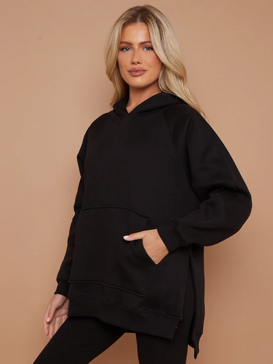 Model wears a black oversized hoodie with font functional pockets and matching  black ribbed leggings. The oversized hoodie has side splits on either side of the jumper.  Model has blonde hair, she stands to the side and tilts her head to face the camera, she slightly smiles. Model has one hand in her pocket and places the other by her side.