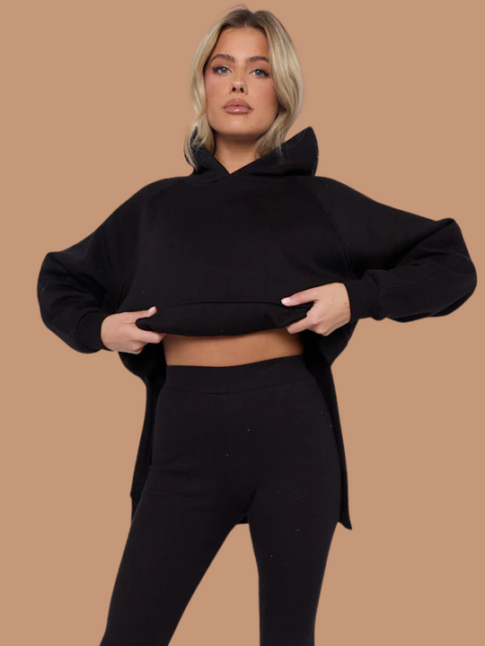 Model wears a black oversized hoodie with front functional pockets and matching  black ribbed leggings. The oversized hoodie has side splits on either side of the jumper.  Model has blonde hair, she holds either side  the hoodie, showing the ribbed leggings.