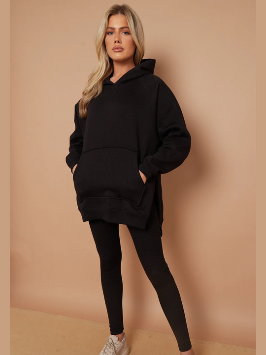 Model wears a black oversized hoodie with front functional pockets and matching  black ribbed leggings. The oversized hoodie has side splits on either side of the jumper.  Model has blonde hair, she faces the caera and places both hands in the hoodies pockets. The full length of the ribbed leggings can also be seen.