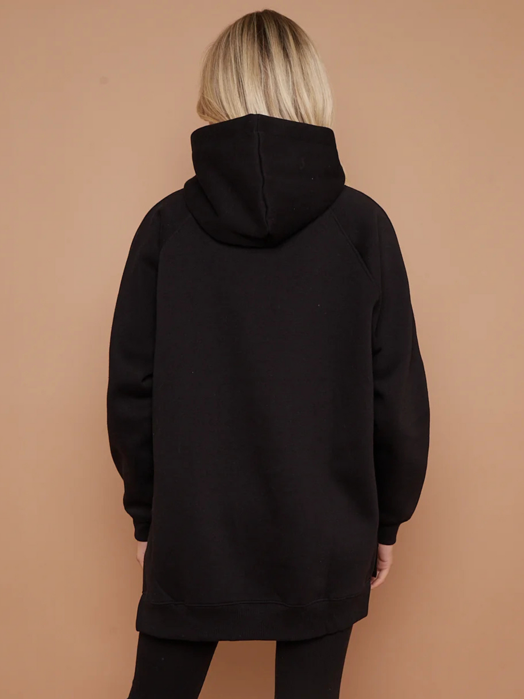 Model wears a black oversized hoodie with front functional pockets and matching  black ribbed leggings. The oversized hoodie has side splits on either side of the jumper.  Model has blonde hair, she stands with her back to the camera, the oversized hoodie length is visible. The hood can be seen. 