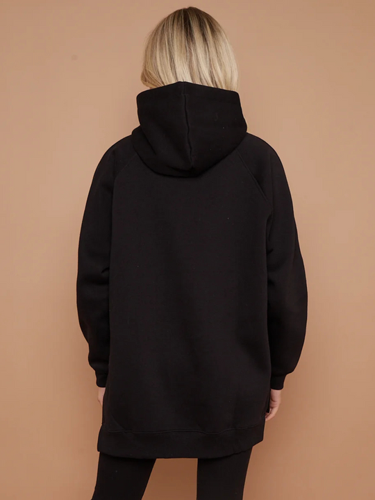 Model wears a black oversized hoodie with front functional pockets and matching  black ribbed leggings. The oversized hoodie has side splits on either side of the jumper.  Model has blonde hair, she stands with her back to the camera, the oversized hoodie length is visible. The hood can be seen. 
