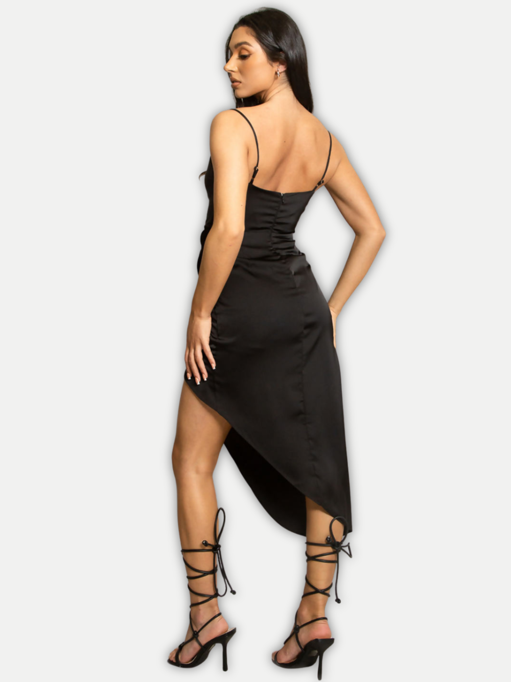 Model stands in front of a light grey background. The model wears a black satin asymmetric midi dress, with a cowl neckline and spaghetti straps. The model has tanned skin and long brown hair, she stands with her back to the camera, the back of the black satin midi dress can be seen, including the concealed zip back fastening. 
