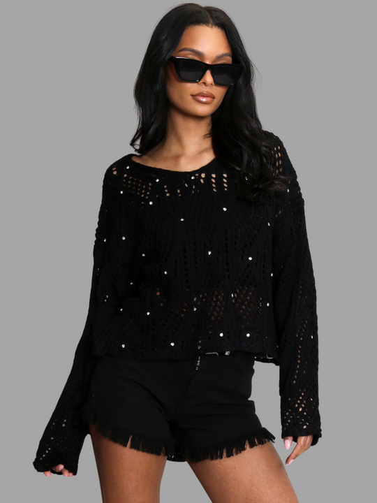 Female model wears a black crochet jumper with long sleeves and front diamante studs. The crochet jumpers has netted pattern with holes visible, the model wears a crop top underneath. The model has light brown skin and black hair, she wears sunglasses, she stands with both hands by her sides. 
