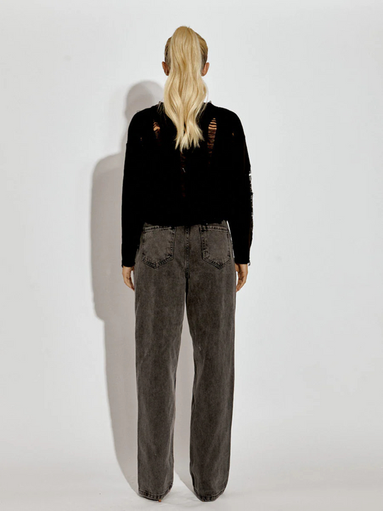 A female model wearing a black  long sleeve distressed style knitted cropped jumper in black color paired with black jeans. The black distressed knitted jumper has distressed panelling  and distressed hems. The model has tanned skin and long blonde hair that is in a ponytail. The model stands with her back to the camers, the back of the jumper including the knitted panelling can be seen. 