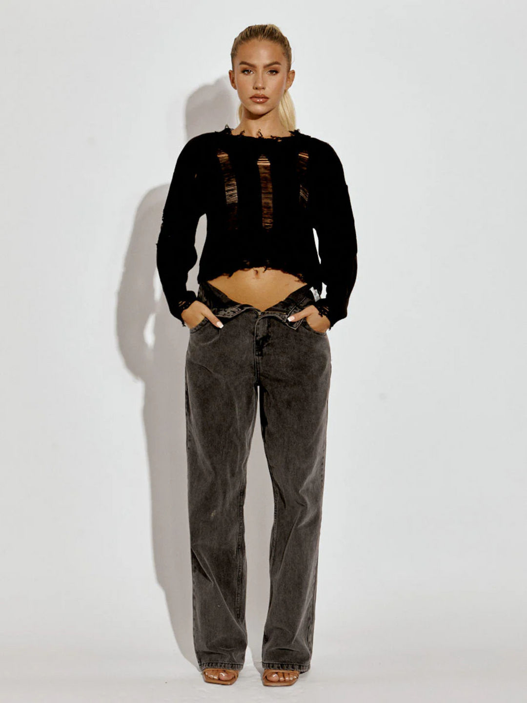 Full length shot. A female model wearing a black  long sleeve distressed style knitted cropped jumper in black color paired with black jeans. The black distressed knitted jumper has distressed panelling  and distressed hems. The model has tanned skin and long blonde hair that is in a ponytail. The model faces the camera, she places her hands in her pockets, the cropped style of the jumper can be seen. 