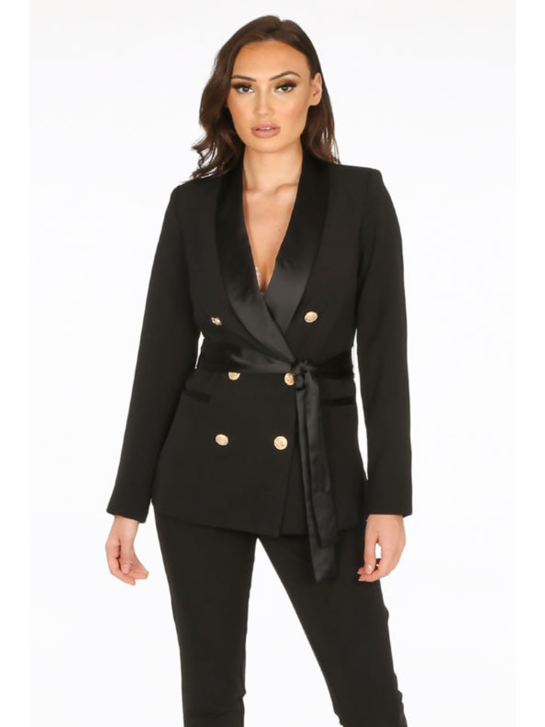 Model stands in front of a plain background. The model wears a luxe tailored black double breasted blazer, with  gold military button detailing and black satin lapel and black satin belt fastening. The model has tanned skin and long brown hair, she faces the amera and wears black trousers.