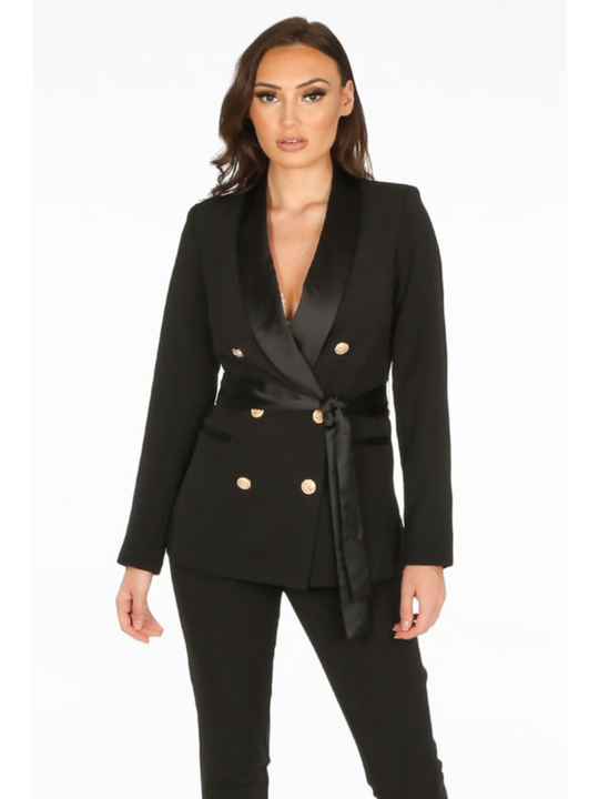 Model stands in front of a plain background. The model wears a luxe tailored black double breasted blazer, with  gold military button detailing and black satin lapel and black satin belt fastening. The model has tanned skin and long brown hair, she faces the amera and wears black trousers.