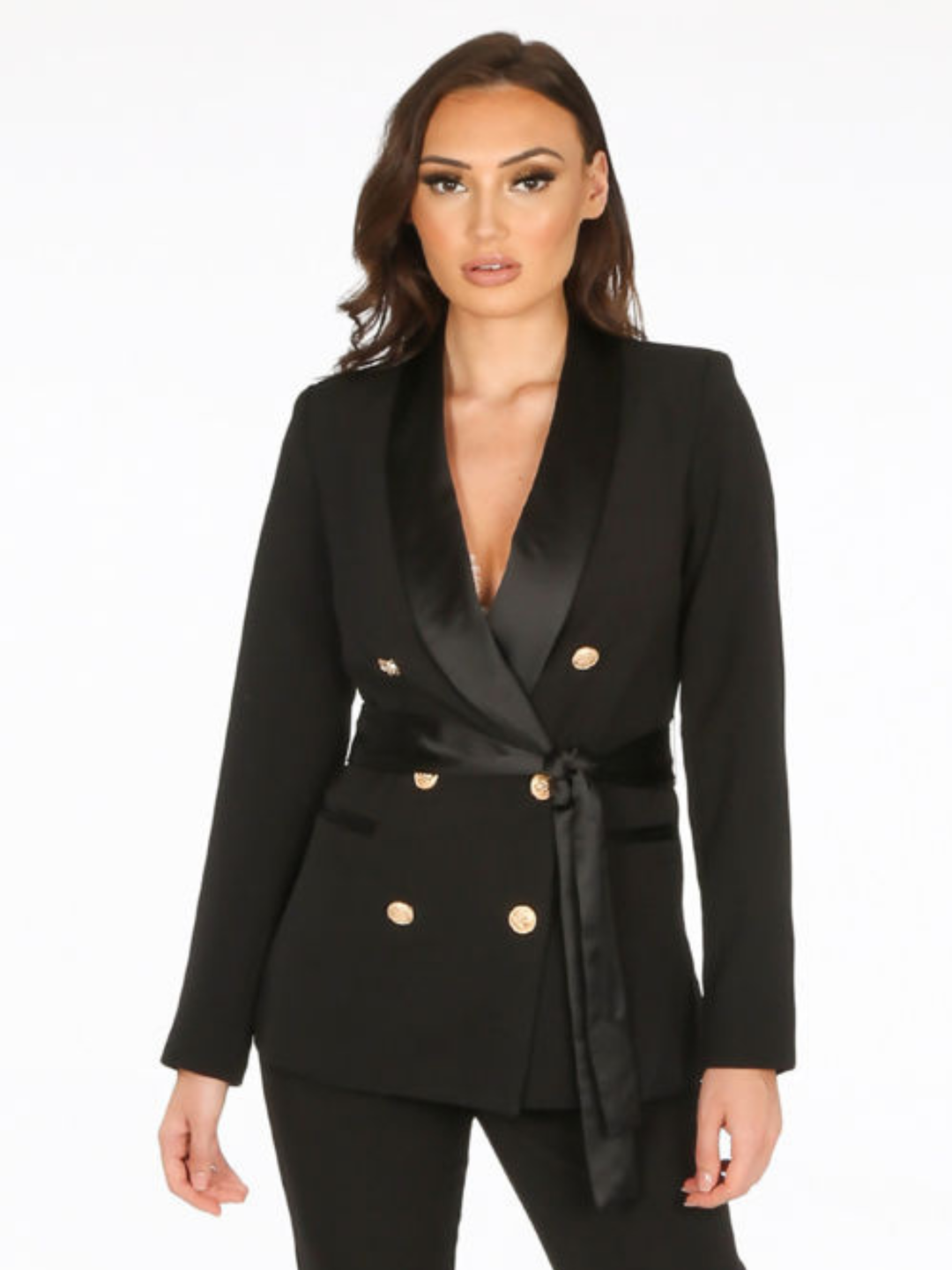 Model stands in front of a plain background. The model wears a luxe tailored black double breasted blazer, with  gold military button detailing and black satin lapel and black satin belt fastening. The model has tanned skin and long brown hair, the model faces the camera and places both hands by her sides.