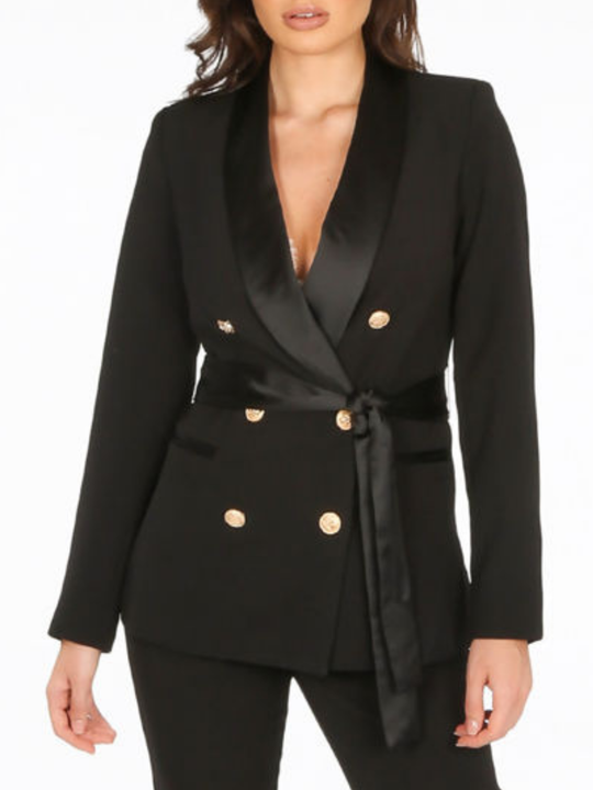 Close up. Model stands in front of a plain background. The model wears a luxe tailored black double breasted blazer, with  gold military button detailing and black satin lapel and black satin belt fastening. 