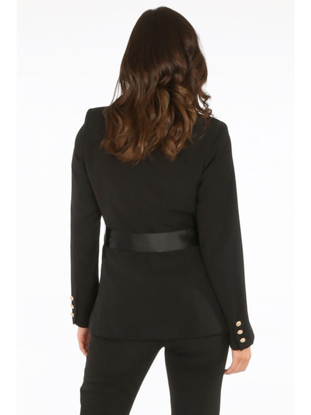Model stands in front of a plain background. The model wears a luxe tailored black double breasted blazer, with  gold military button detailing and black satin lapel and black satin belt fastening. The model has tanned skin and long brown hair, she stands with her back to the camera, the back of the blazer can be seen, including the gold military button sleeve cuffed design.
