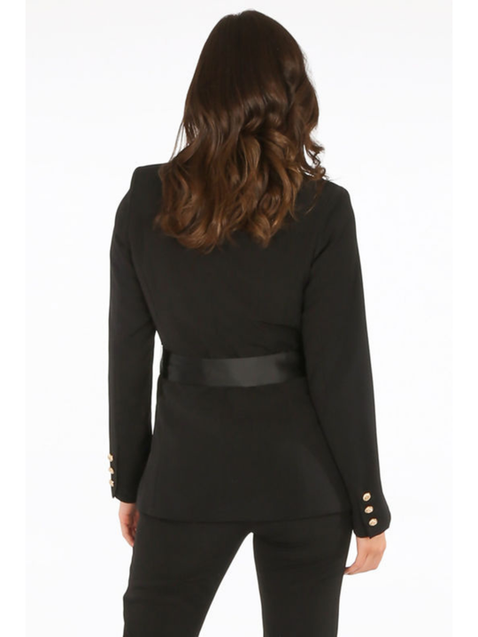 Model stands in front of a plain background. The model wears a luxe tailored black double breasted blazer, with  gold military button detailing and black satin lapel and black satin belt fastening. The model has tanned skin and long brown hair, she stands with her back to the camera, the back of the blazer can be seen, including the gold military button sleeve cuffed design.