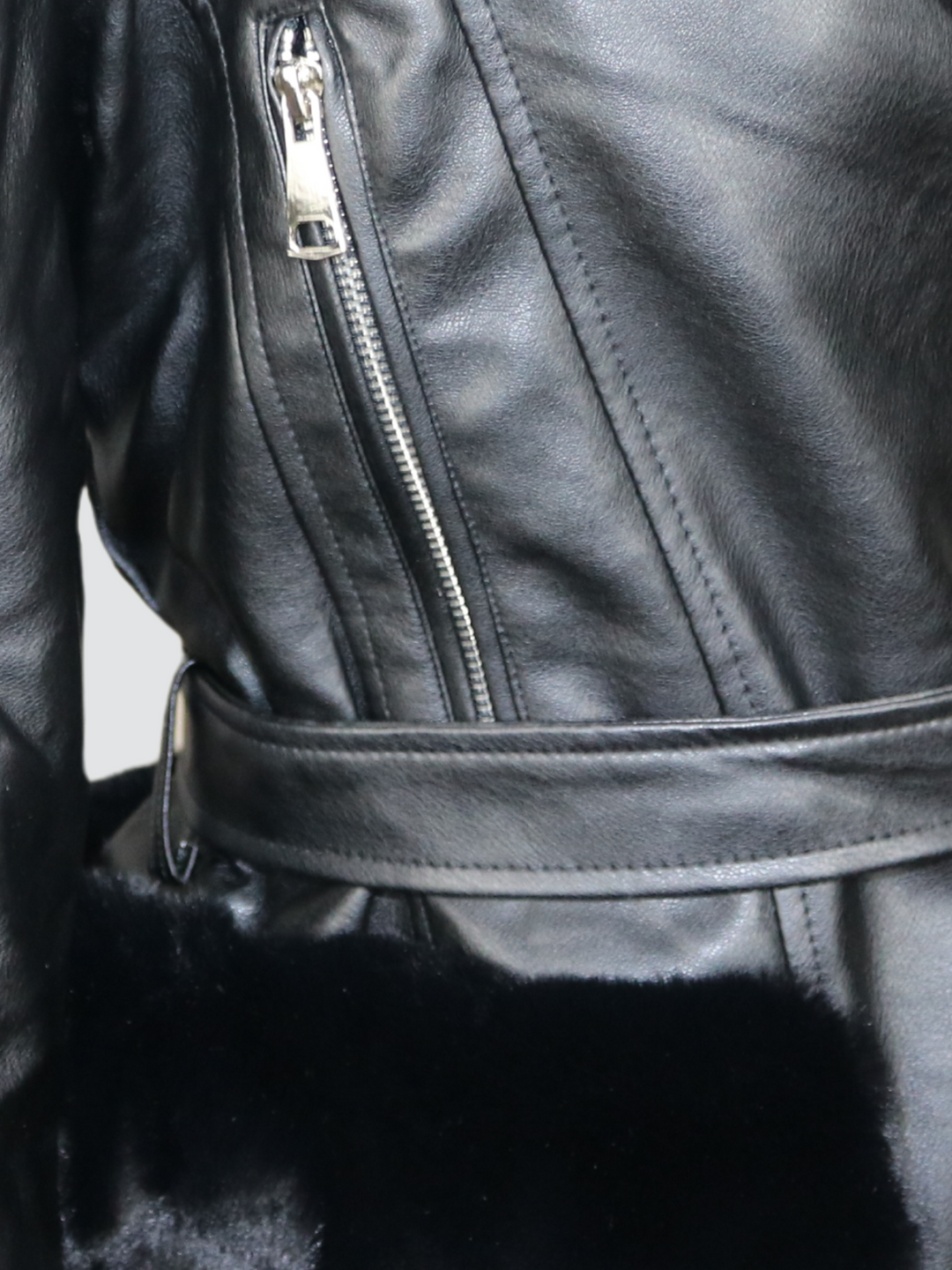 Ghost manequin wears a biker faux leather jacket with faux fur trim. The jacket has leather panelled detailing at the sleeves and exposed asymmetric chunky zip fastening. The faux leather jacket has a removable faux  belted waist.  A close up of the trunk of jacket can be seen, including a belted waist, faux fur trim and exposed chunky zip fastening. 