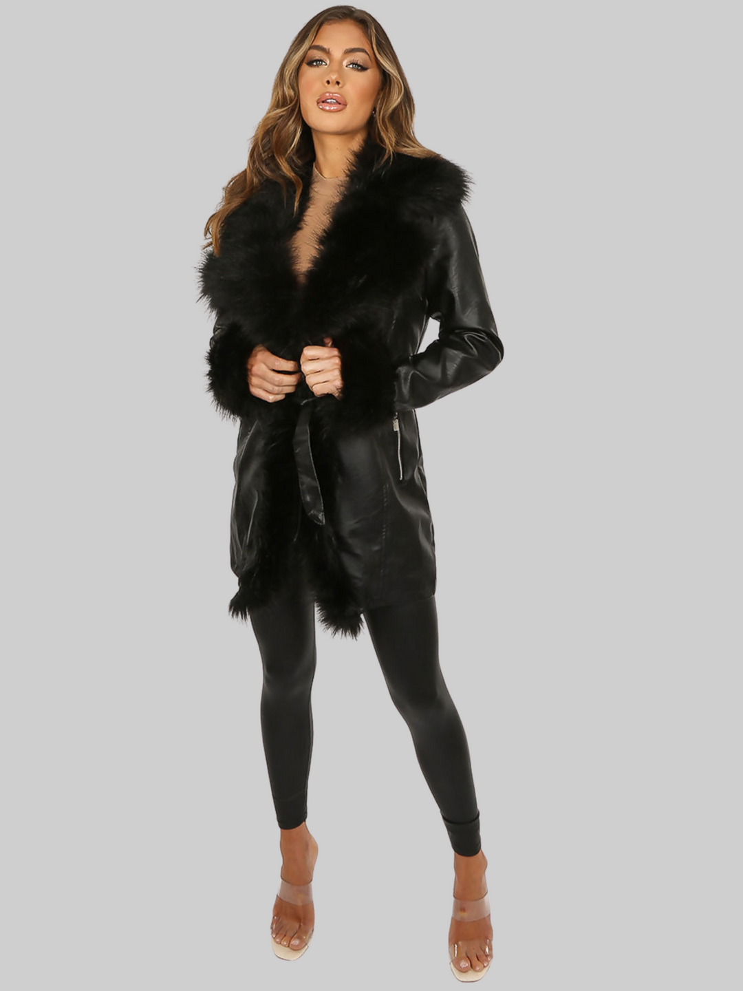 Full length shot. Model wears a black faux leather  coat with a longline silhouette. The coat has a faux fur ttrim and faux fur cuffs, there is a removable faux leather belt at the waist and side zip pockets.  Model faces the camera, she has both hands in front of her waist and wears pu leggings and clear heels. 