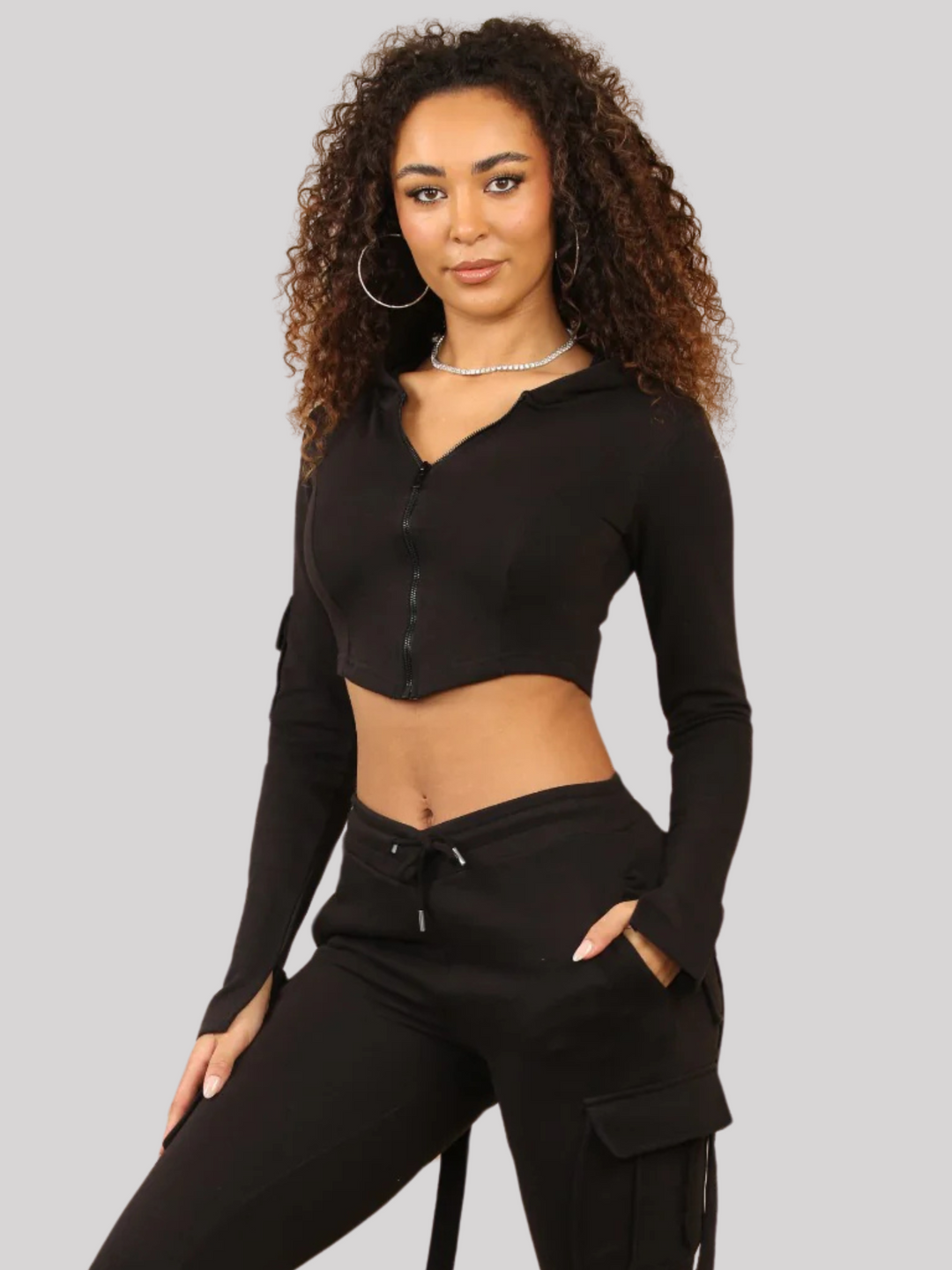 Model wears a Black Cropped Top Hoodie With Zip Front Fastening and Fitted Chargo Jogger Set. The cargo jogger has pockets and one pocket on the long sleeve hoodie. Model has light brown skin, curly textured hair and stares into the camera. She stands with her leg to the side ans places one hand in her pocket and the other on her leg. 
