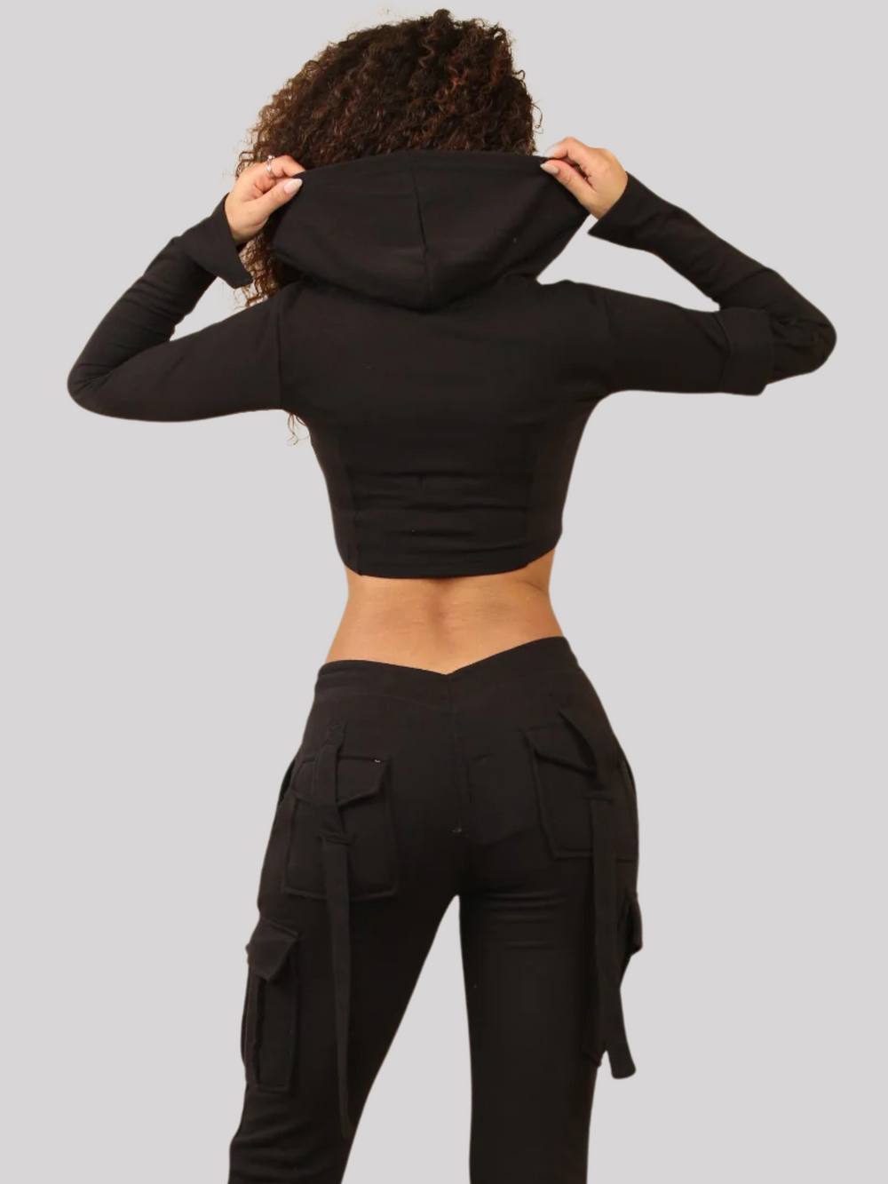 Model wears a Black Cropped Top Hoodie With Zip Front Fastening and Fitted Chargo Jogger Set. The cargo jogger has pockets and one pocket on the long sleeve hoodie. Model has light brown skin, curly textured hair, she stands with her back to the camera, the hooded crop top and back of the cargo joggers can be see. 