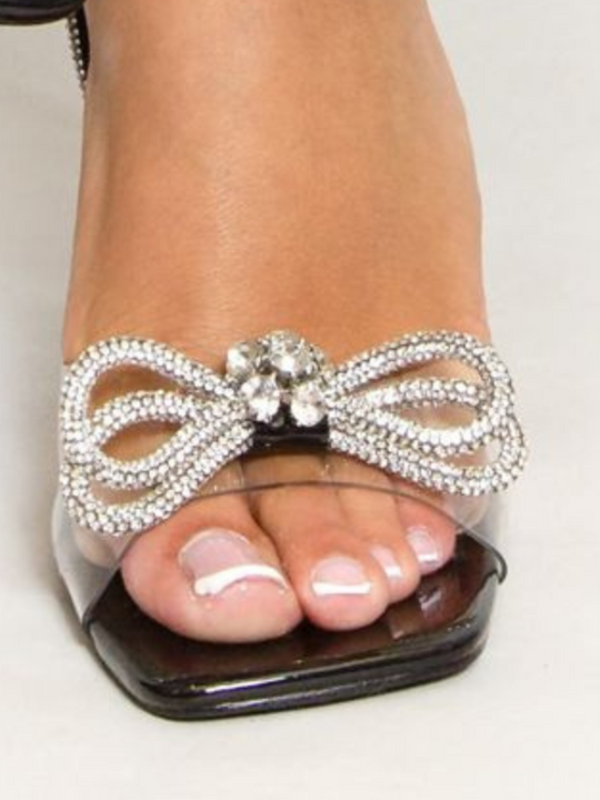 Close up of the diamante bow front design. Model wears Model wears an open toe shoe with a front perspex strap, diamante bow front design and diamante ankle strap. 