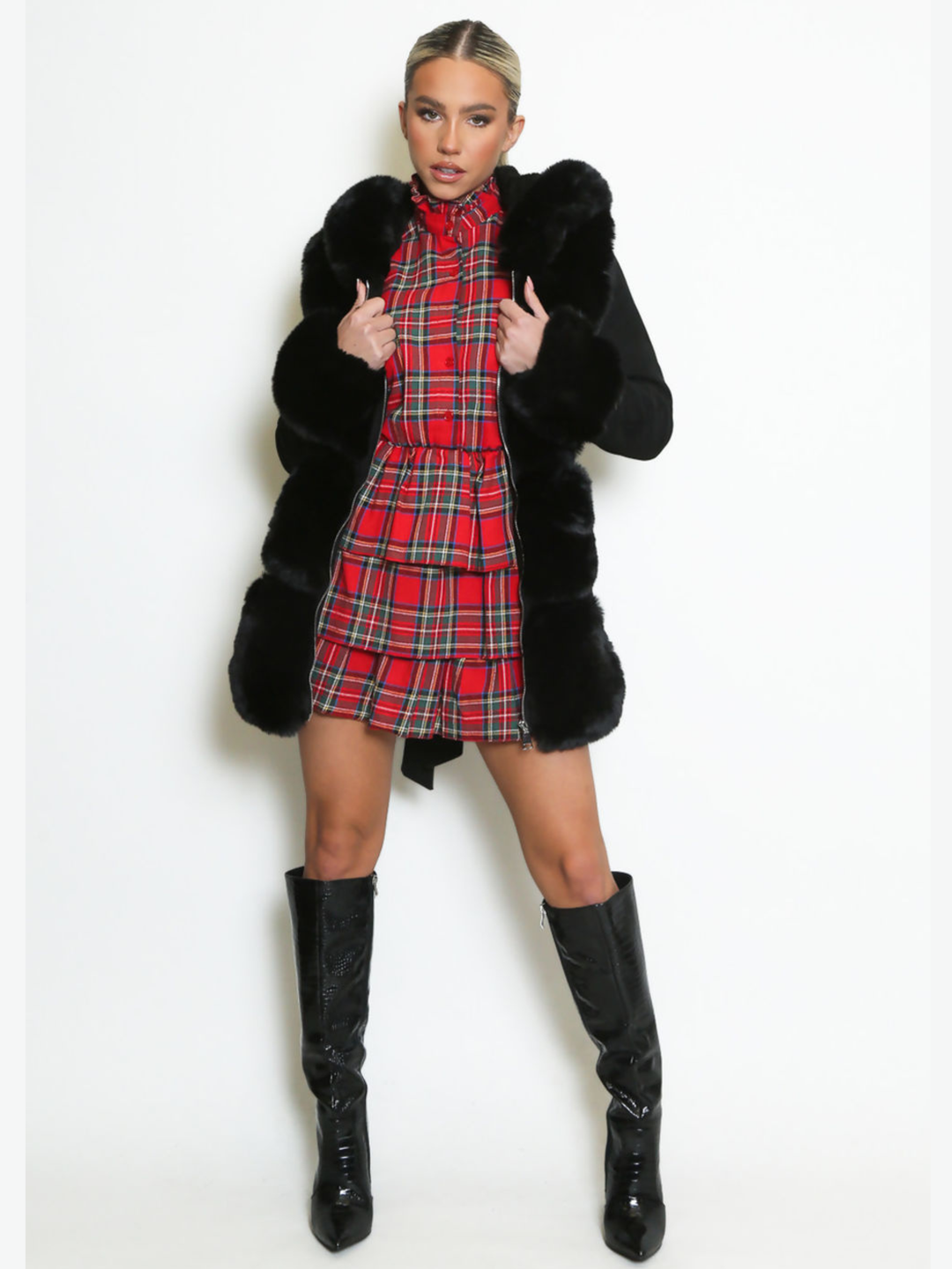 Full length shot. Female model stands in front of a plain background, she wears a longline jacket with faux fur front panelling, faux fur sleeved cuffs,  exposed zip front fastening and a belted waist. The model has tanned skin and blonde hair that has been slicked back into a ponytail, she stands facing forwards with the coat open, wearing a tartan mini dress underneath, the faux fur panel can be seen, as well as the faux fur sleeved cuffs, she holds either side of the open coat and wears black boots. 