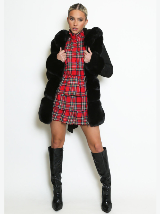 Full length shot. Female model stands in front of a plain background, she wears a longline jacket with faux fur front panelling, faux fur sleeved cuffs,  exposed zip front fastening and a belted waist. The model has tanned skin and blonde hair that has been slicked back into a ponytail, she stands facing forwards with the coat open, wearing a tartan mini dress underneath, the faux fur panel can be seen, as well as the faux fur sleeved cuffs, she holds either side of the open coat and wears black boots. 