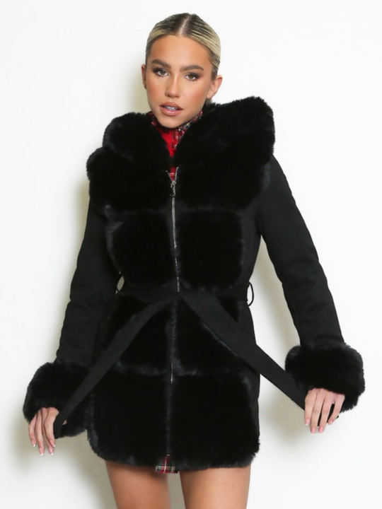 Female model stands in front of a plain background, she wears a longline jacket with faux fur front panelling, faux fur sleeved cuffs,  exposed zip front fastening and a belted waist. The model has tanned skin and blonde hair that has been slicked back into a ponytail, she stares into the camera and holds the waist belt on either side.  
