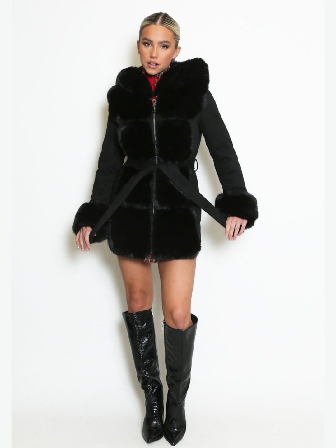 Full length shot. Female model stands in front of a plain background, she wears a longline jacket with faux fur front panelling, faux fur sleeved cuffs,  exposed zip front fastening and a belted waist. The model has tanned skin and blonde hair that has been slicked back into a ponytail, she stares into the camera and holds the waist belt on either side.  The model wears black knee high boots.