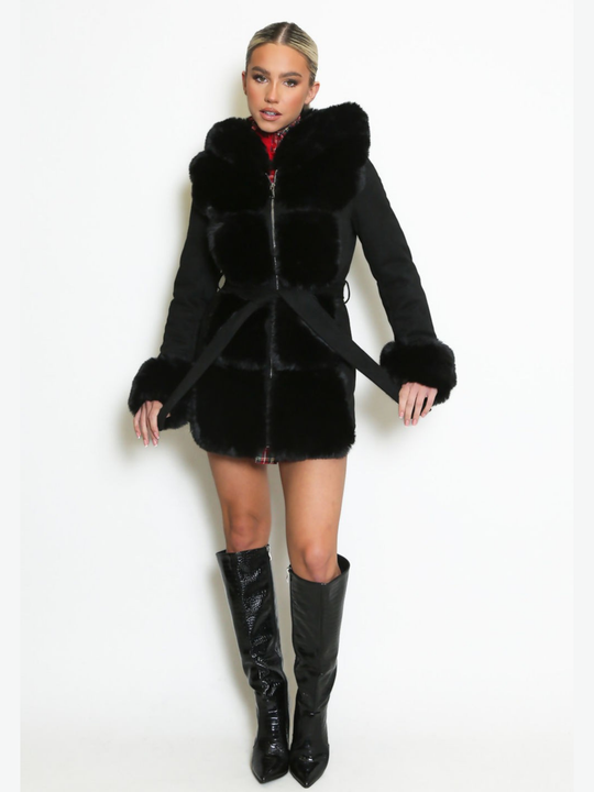 Full length shot. Female model stands in front of a plain background, she wears a longline jacket with faux fur front panelling, faux fur sleeved cuffs,  exposed zip front fastening and a belted waist. The model has tanned skin and blonde hair that has been slicked back into a ponytail, she stares into the camera and holds the waist belt on either side.  The model wears black knee high boots.