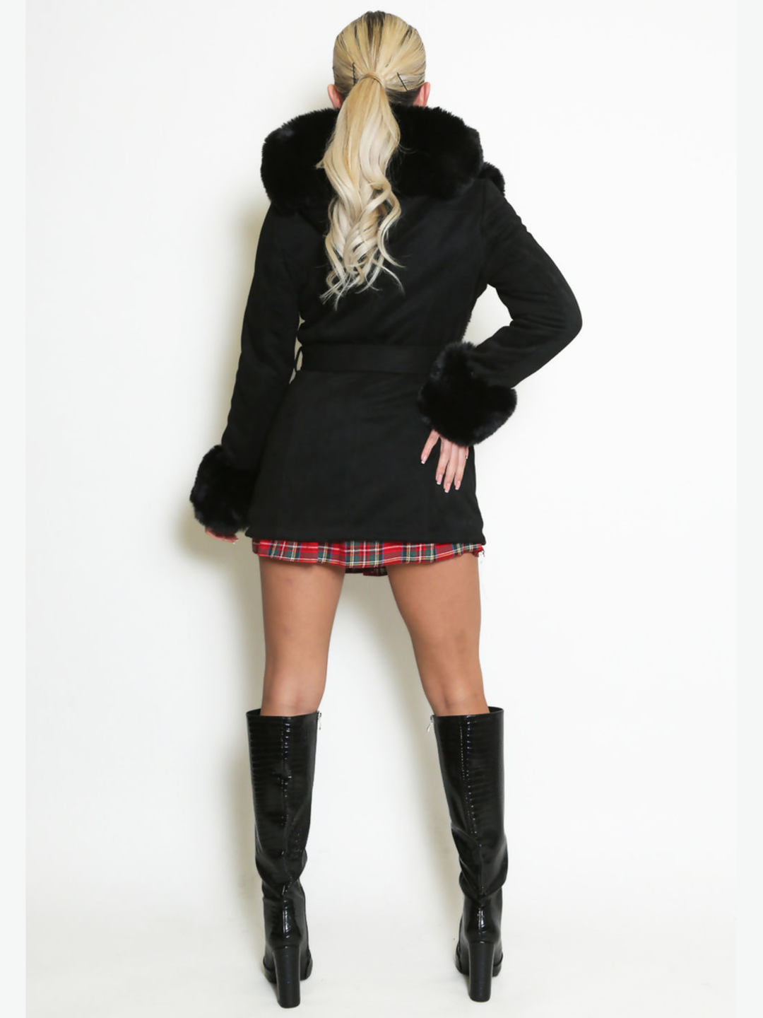 Full length shot. Female model stands in front of a plain background, she wears a longline jacket with faux fur front panelling, faux fur sleeved cuffs,  exposed zip front fastening and a belted waist. The model has tanned skin and blonde hair that has been slicked back into a ponytail, she stands with her back to the camera, placing one hand on her hip and the other by her side, the back of the coat can be seen including the dented waist and four fur cuffs, she wears black knee high boots. 