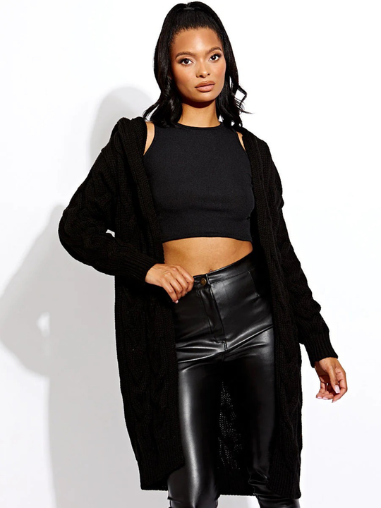 Female model stands in front of a plain background. The female model wears a long open cable knit Cardin in black. The cable knit cardigan has long sleeves. The model has light brown skin and long black hair, she faces the camera, placing one hand on her trouser waist and the other by her side. 