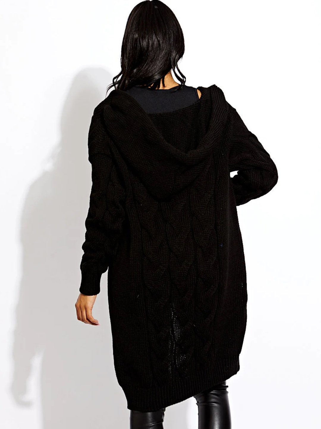 Female model stands in front of a plain background. The female model wears a long open cable knit Cardin in black. The cable knit cardigan has long sleeves. The model has light brown skin and long black hair,  she stands with her back to the camera, the back of the cardigan can be seen, including the cardigan's hood. 