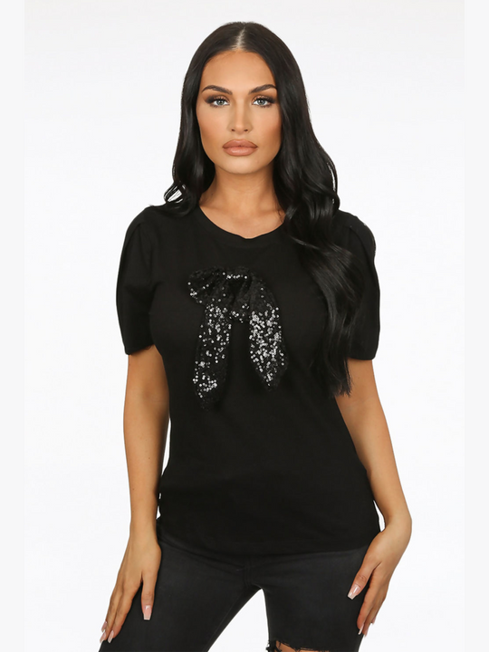 Female model wears a luxe black t shirt with a rounded neck amd 3/4 sleeves, with a large black sequin bow. The model faces the camera in front of  a plain background,  she has tanned skin and long black hair. The model places both hands by her sides. 