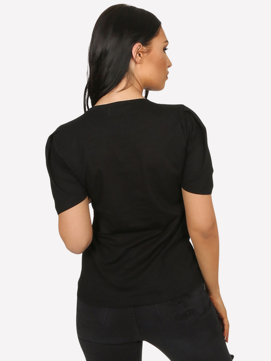 Female model wears a luxe black t shirt with a rounded neck amd 3/4 sleeves, with a large black sequin bow. The model  has tanned skin and long black hair, she stands with her back to the camera, the plain back of the t shirt can be seen.  