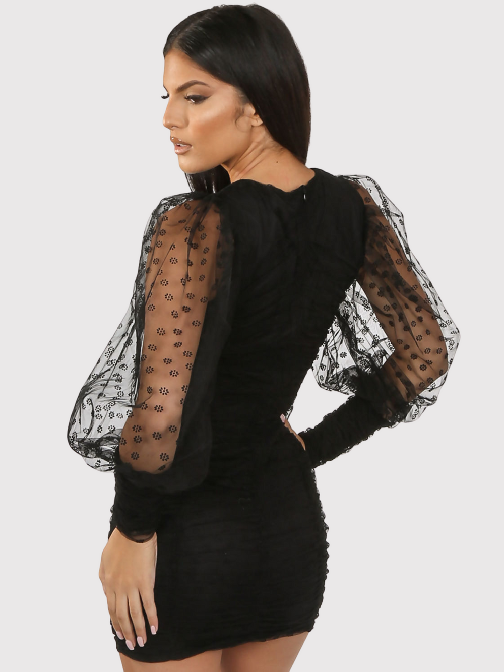 Female model wears a Black mini dress with sheer long sleeves with rounded neck and pleated overlay.  The model has tanned skin and long black hair, she stands with her back to the camera, the back of the dress can be seen, as well as the sheer sleeves. 