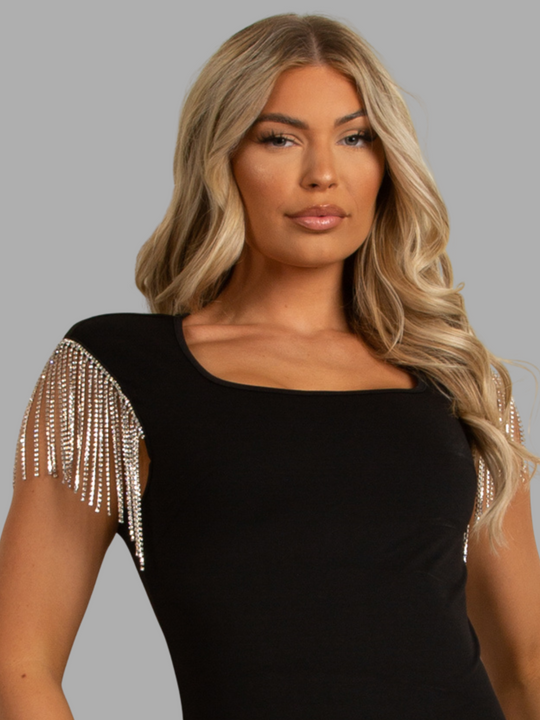 Female model stands in front of a grey plain background. The female model wears a luxe black mini dress, with square neck design and short diamante tasseled sleeves.  The model has tanned skin and blonde hair, she faces forward, the side of the diamante tasseled sleeve can be seen. 