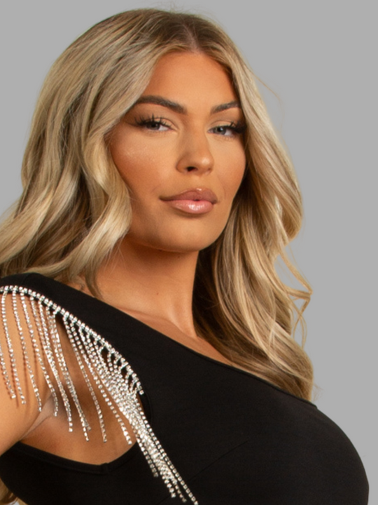 Female model stands in front of a grey plain background. The female model wears a luxe black mini dress, with square neck design and short diamante tasseled sleeves.  The model has tanned skin and blonde hair, she looks into the camera, the diamante tasseled sleeves can be seen. 