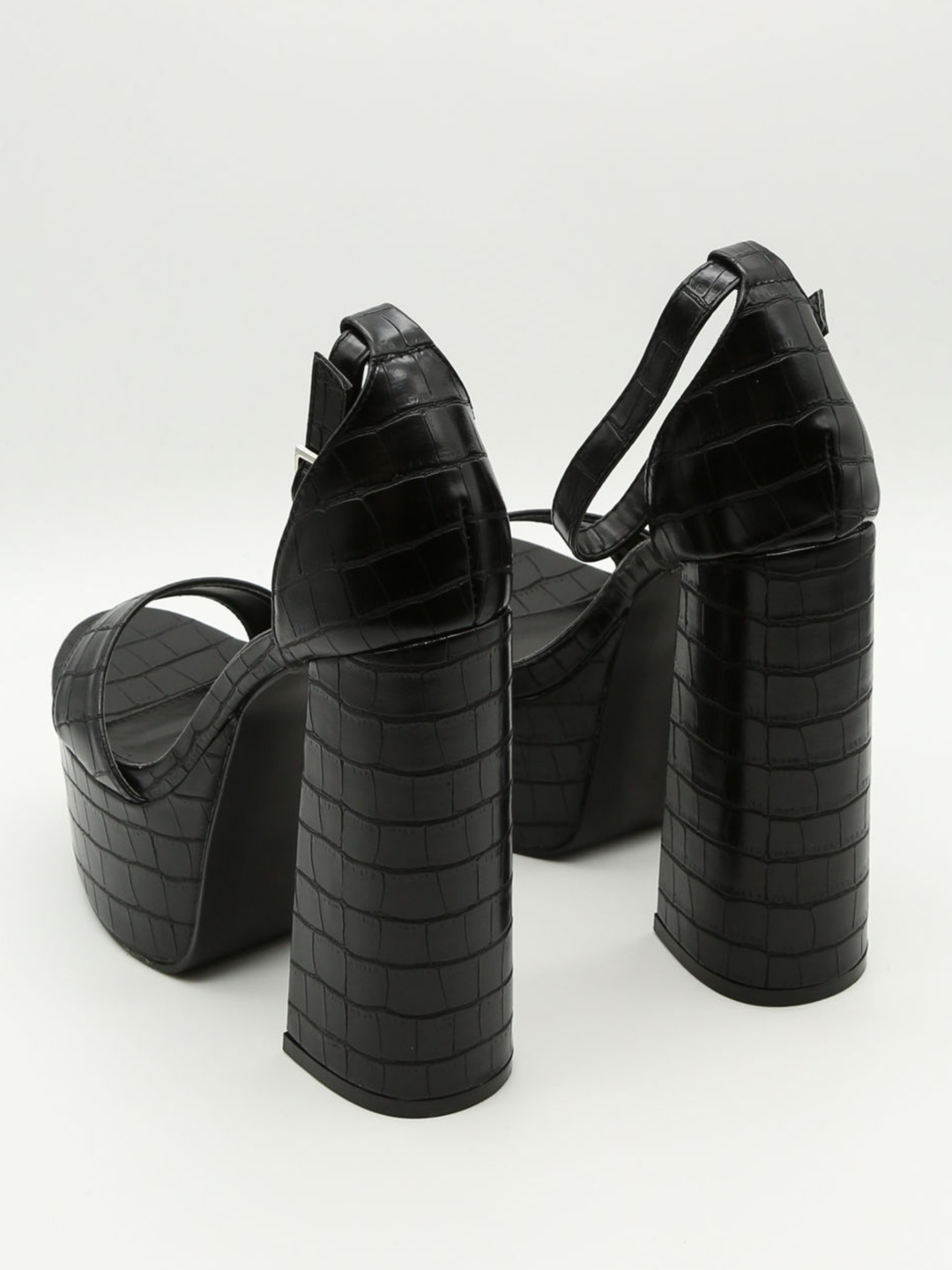 A croc chunky shoe is placed in front of a plain background. The model wears  a black platform shoe with chunky heels and adjustable ankle strap fastening.  The shoe is placed  with the back side of the shoe to the camera, showing the chunky heels and the croc faux leather design. 
