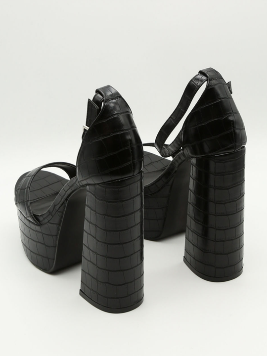 A croc chunky shoe is placed in front of a plain background. The model wears  a black platform shoe with chunky heels and adjustable ankle strap fastening.  The shoe is placed  with the back side of the shoe to the camera, showing the chunky heels and the croc faux leather design. 
