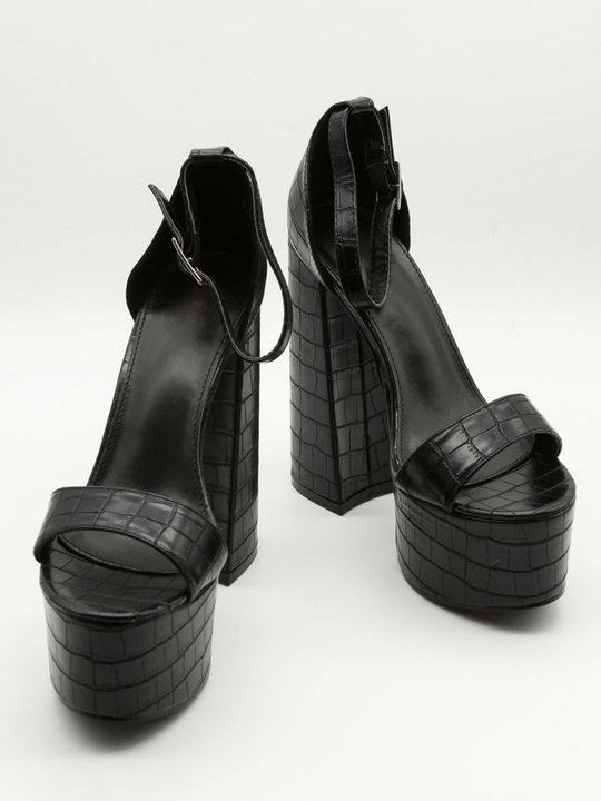 A croc chunky shoe is placed in front of a plain background. The model wears  a black platform shoe with chunky heels and adjustable ankle strap fastening.  The shoe is placed  with the front of the shoe, showing the front foot strap and adjustable ankle straps.