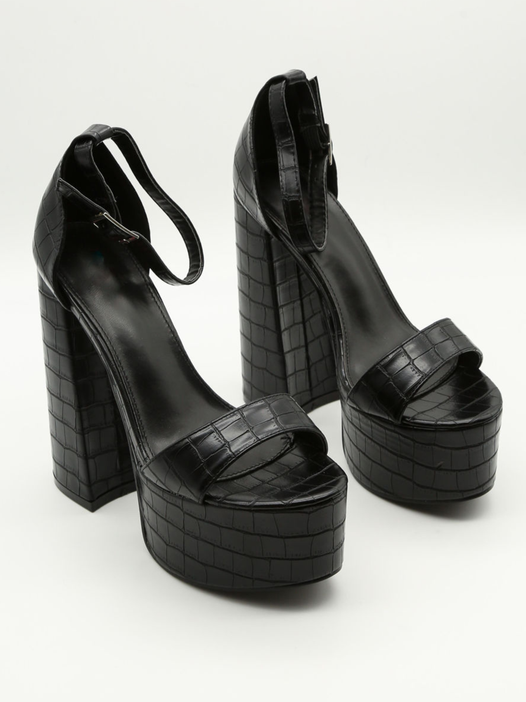 A croc chunky shoe is placed in front of a plain background. The model wears  a black platform shoe with chunky heels and adjustable ankle strap fastening.  The shoe is placed at a slight angle, the front and the side of the shoe can be seen. 